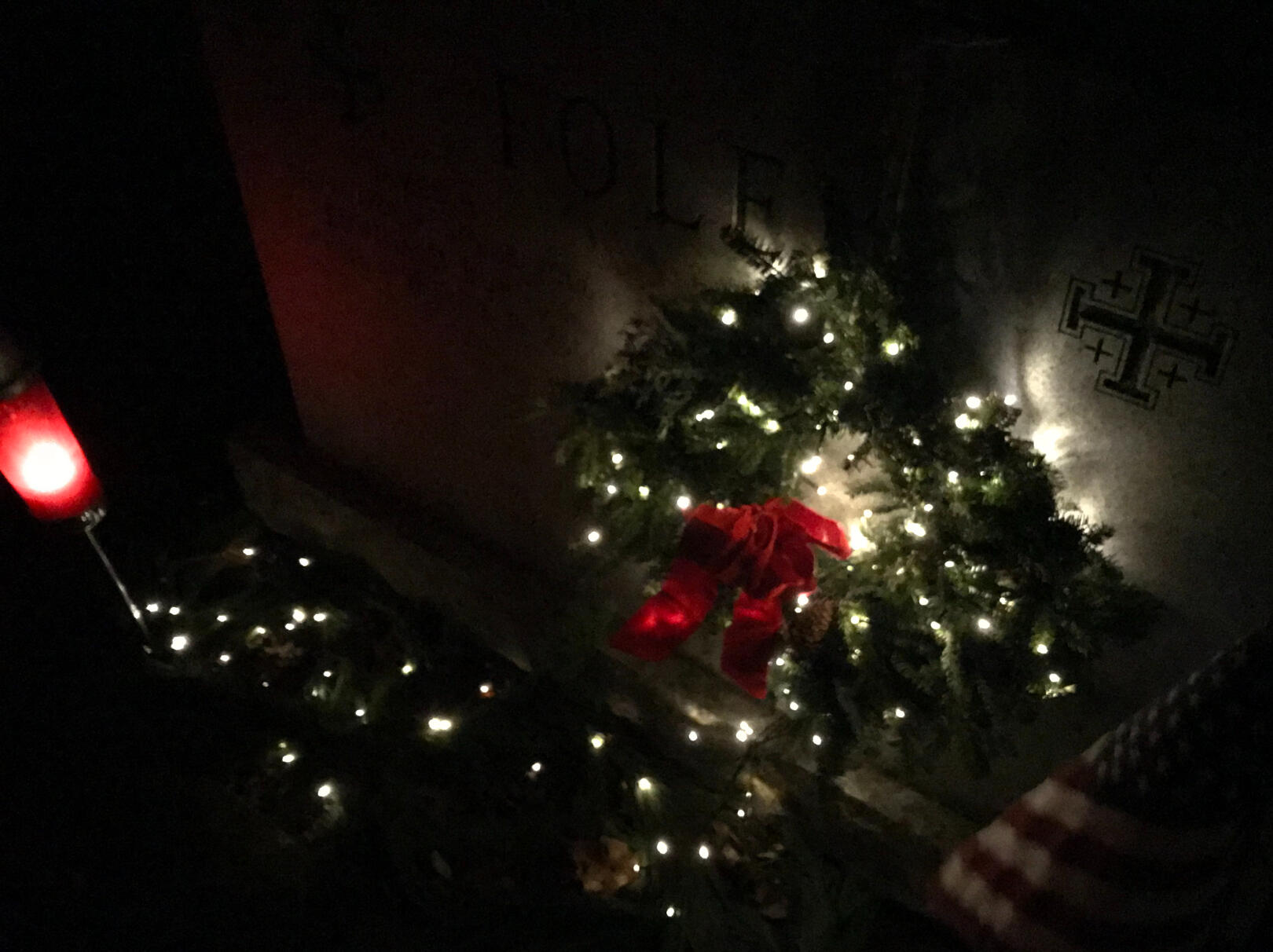 A grave site at night decorated with a lit Christmas wreath, warm white lights, and a red velvet bow